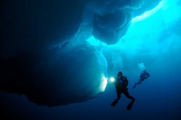 Scuba divers swimming under ice in deep blue water