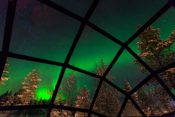View from inside a glass igloo bedroom looking up at green aurora in the night sky