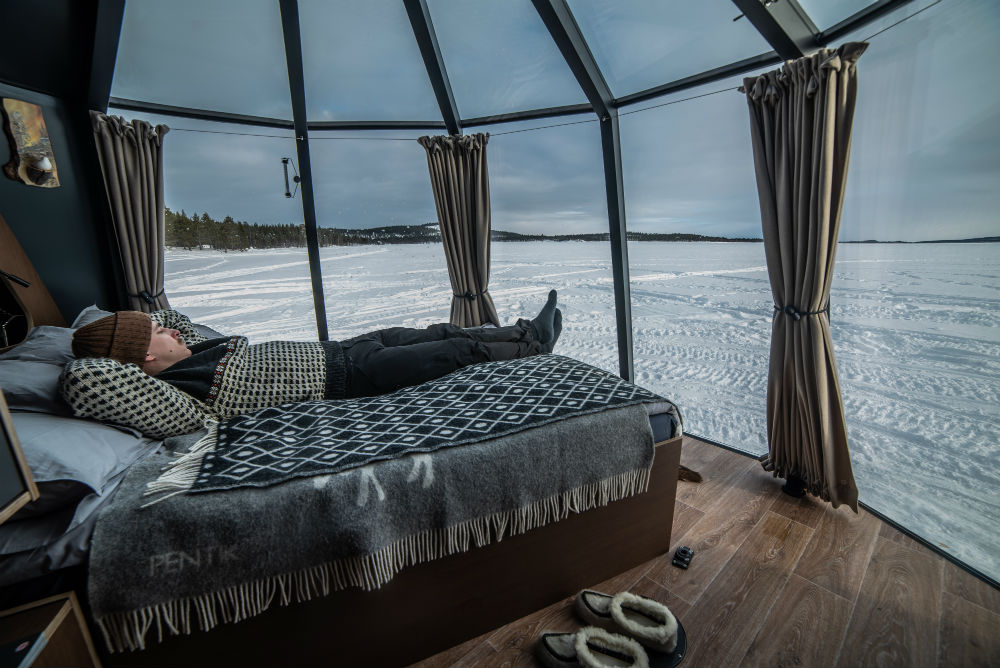 person lying on the bed in an aurora hut overlooking the frozen lake