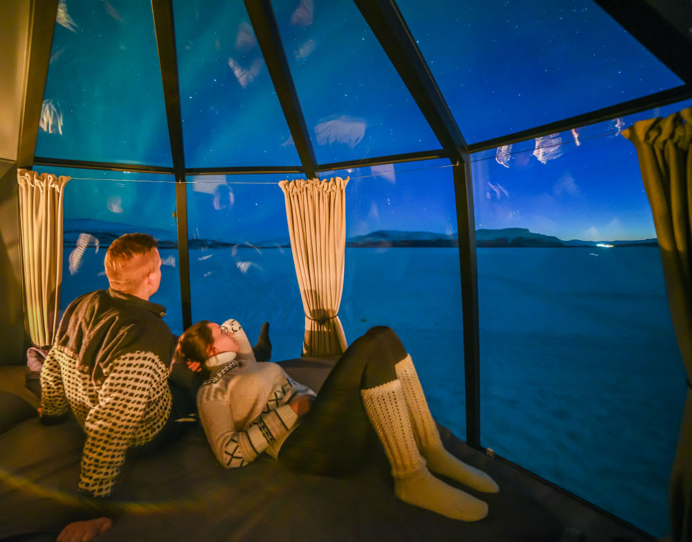 2 people sat on the bed in aurora hut overlooking the frozen lake at night