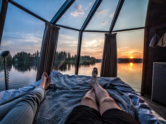 View from a glass-roof cabin bed with two peopleโs legs stretched out, watching a sunset over a calm forest lake.