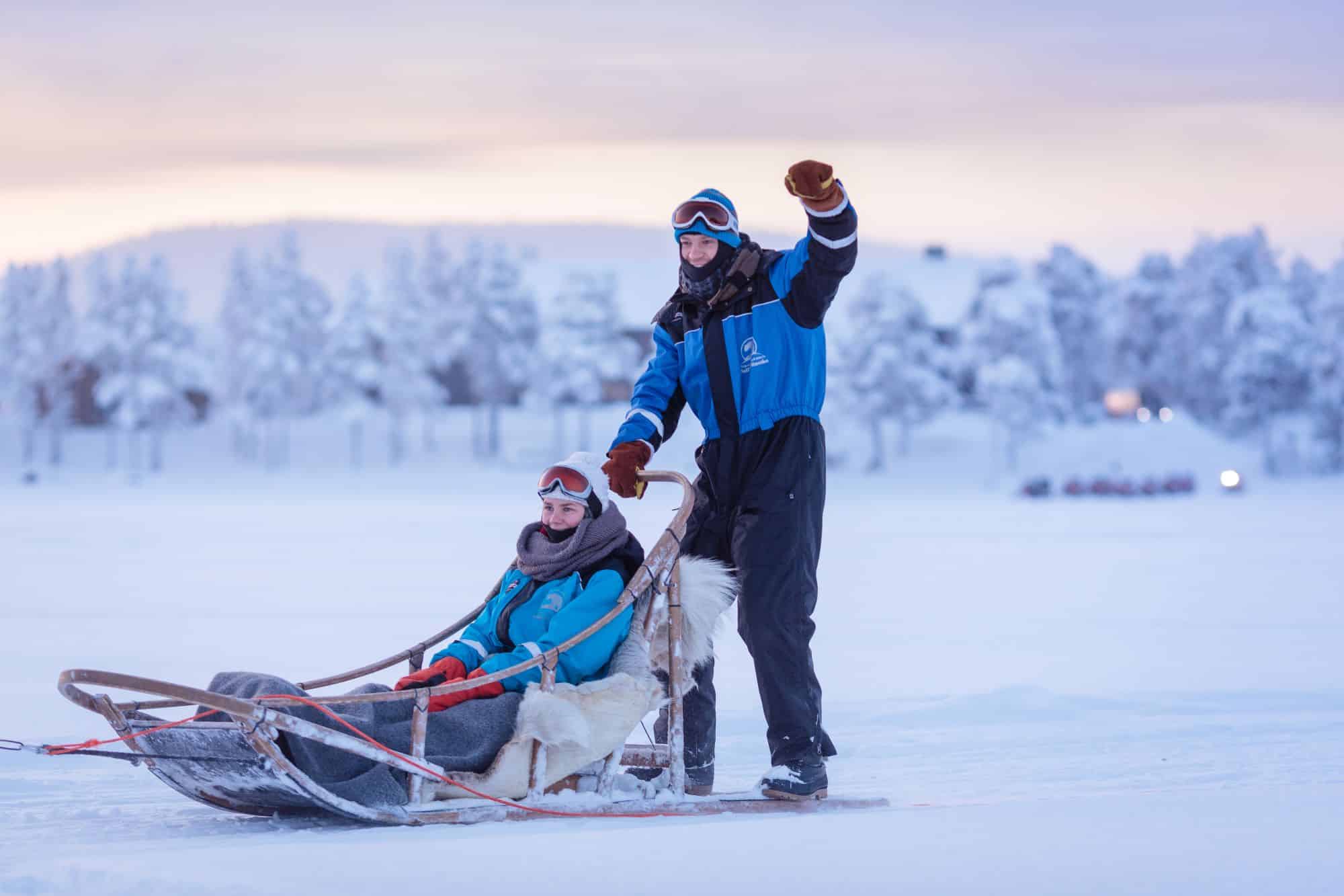 woman sat in husky sled and man on the back driving the huskies