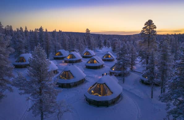 snow covered glass igloo cabins in the forest with sunset behind
