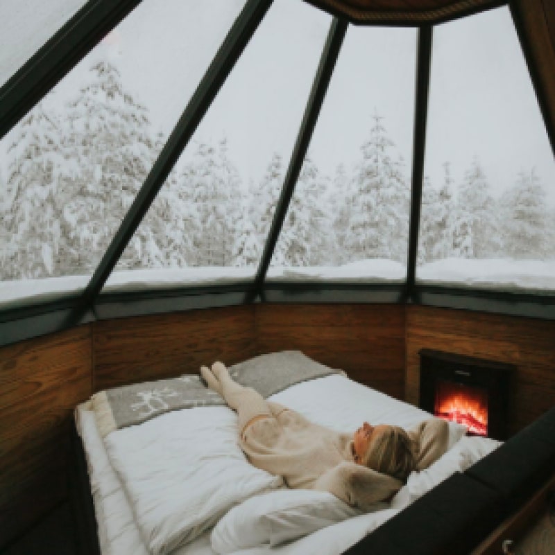Woman relaxing on a bed inside a glass-roof igloo with snowy forest outside