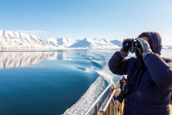 Person in a winter parka using binoculars on a boat, with snowy Arctic mountains reflected in calm blue water.